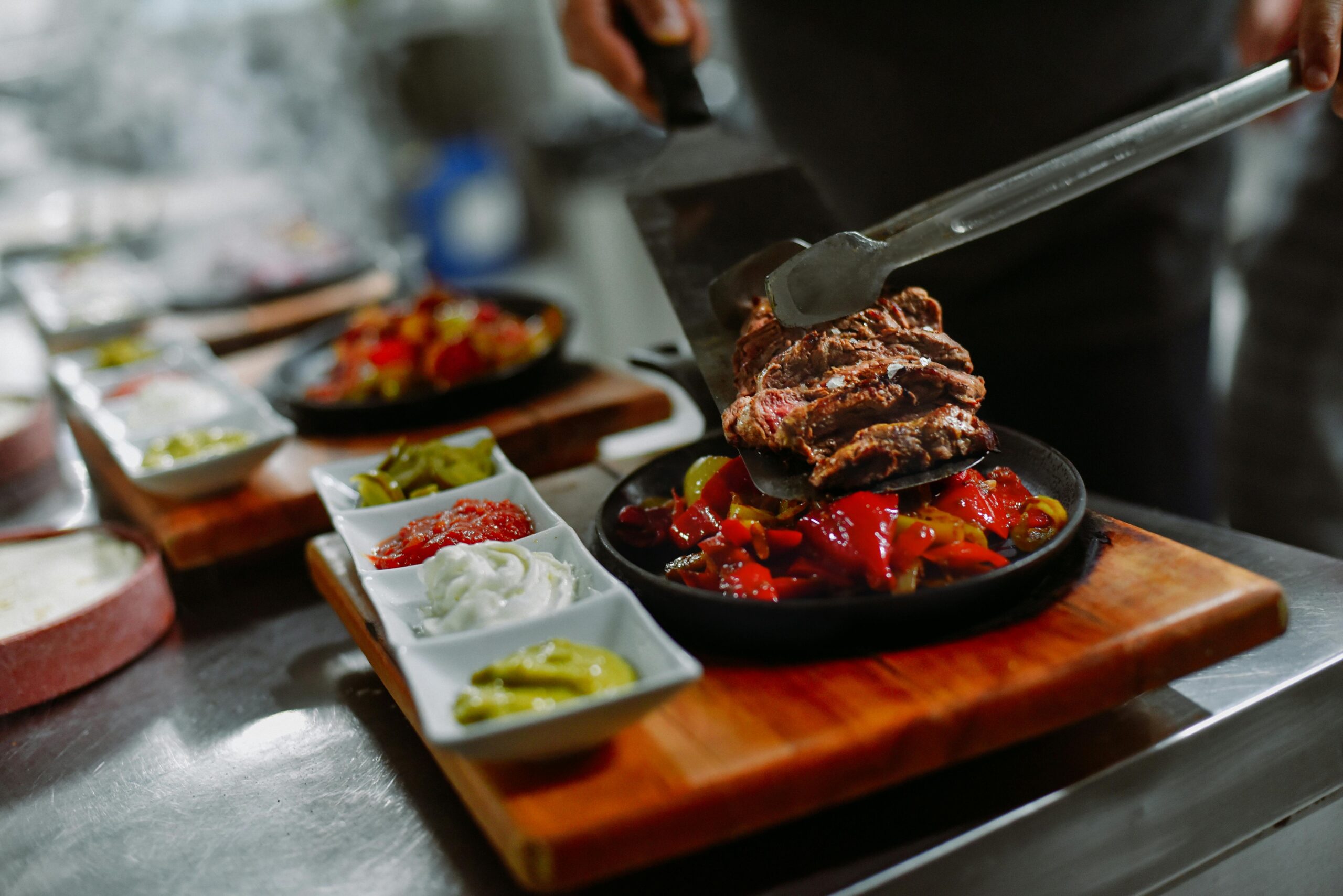 Close-up of a chef preparing grilled steak with vegetables and sauces on a wooden board in a restaurant kitchen.